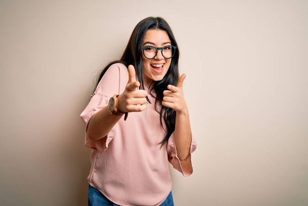 Jeune femme brune élégante portant des lunettes sur fond isolé pointant les doigts vers la caméra avec un visage heureux et drôle. Bonne énergie et vibes. - Photo, image
