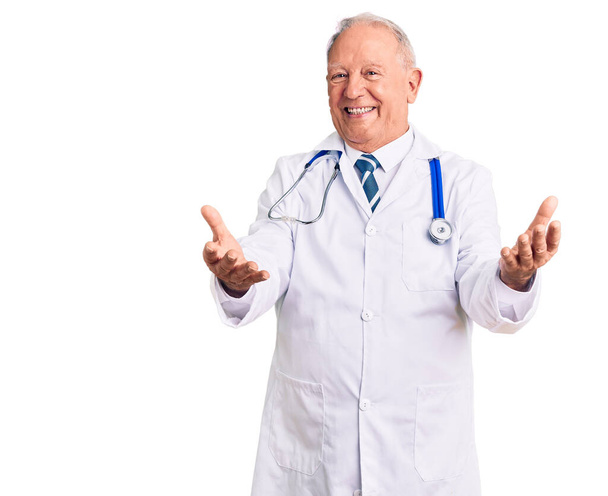 Älterer gutaussehender grauhaariger Mann mit Arztkittel und Stethoskop lächelt fröhlich und bietet Hände an, die Hilfe und Akzeptanz geben.  - Foto, Bild