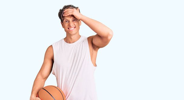 Młody przystojny mężczyzna trzyma piłkę do koszykówki zestresowany i sfrustrowany ręką na głowie, zaskoczony i zły twarz  - Zdjęcie, obraz