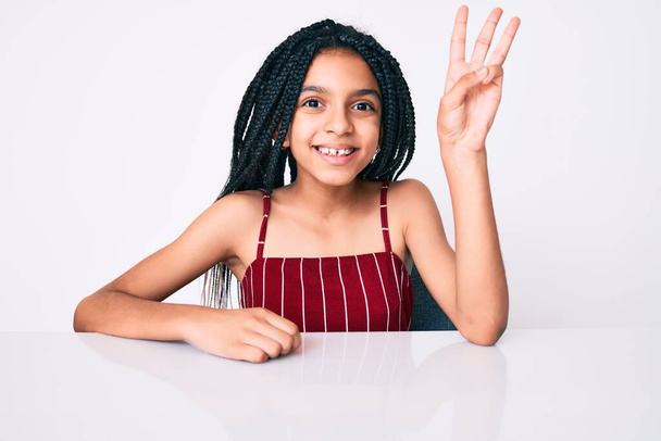 Giovane bambina afroamericana con trecce che indossa abiti casual seduta sul tavolo mostrando e indicando con le dita il numero tre mentre sorride fiducioso e felice.  - Foto, immagini