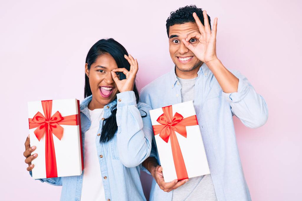 Hermosa pareja latina joven sosteniendo regalo para la celebración del aniversario sonriendo feliz haciendo signo de ok con la mano en el ojo mirando a través de los dedos
  - Foto, Imagen