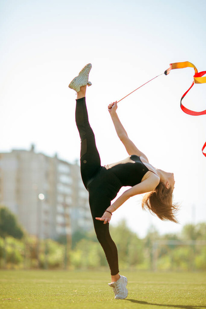 mladá dobře vypadající dívka v černošky sportovní oblečení dělá gymnastika splits v parku drží gymnastický pruh - Fotografie, Obrázek