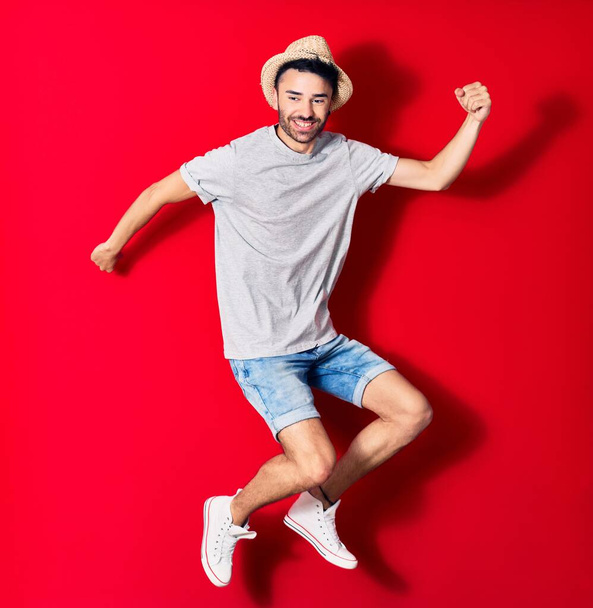 Young handsome hispanic man wearing casual clothes and summer hat smiling happy. Jumping with smile on face celebrating with fists up over isolated red background. - Φωτογραφία, εικόνα