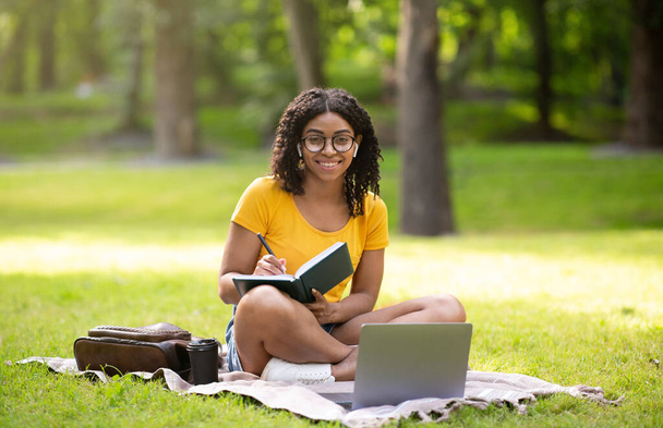 Millennial Afroamerykanka studiuje zdalnie na laptopie, zapisując informacje w notatniku, na zewnątrz - Zdjęcie, obraz
