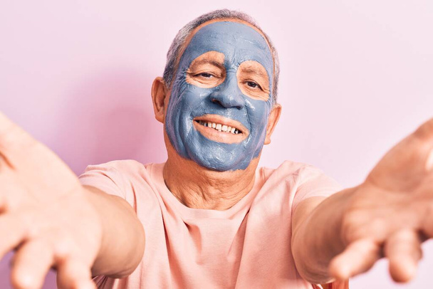 Homme âgé aux cheveux gris portant un masque de boue regardant la caméra souriant à bras ouverts pour un câlin. expression joyeuse embrassant le bonheur.  - Photo, image