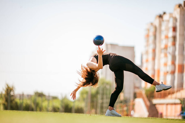 νέα όμορφη κοπέλα σε μαύρο αθλητικό ντύσιμο κάνει γυμναστική handstand στις σχισμές στο πάρκο με μπάλα - Φωτογραφία, εικόνα