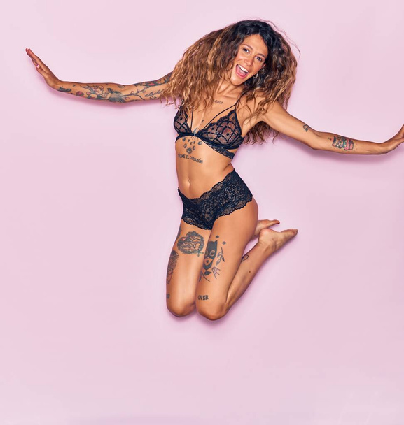 Young beautiful curly woman with tattoo wearing lingerie smiling happy. Jumping with smile on face and arms opened over isolated pink background - Photo, Image