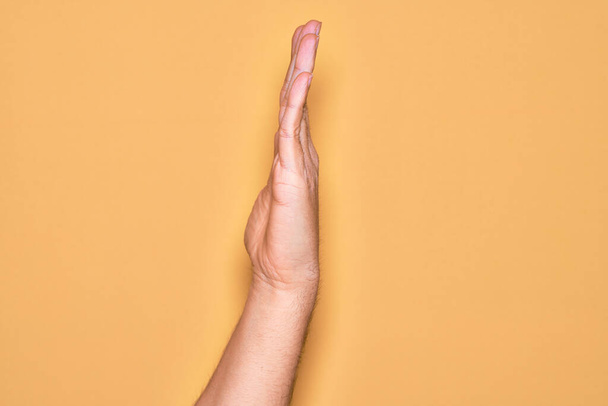 Mano de joven caucásico mostrando los dedos sobre el fondo amarillo aislado mostrando el lado de la mano estirada, empujando y haciendo gesto de parada - Foto, imagen