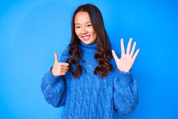 Junge schöne Chinesin trägt lässigen Winterpullover und zeigt mit Fingern Nummer sechs nach oben, während sie selbstbewusst und glücklich lächelt.  - Foto, Bild