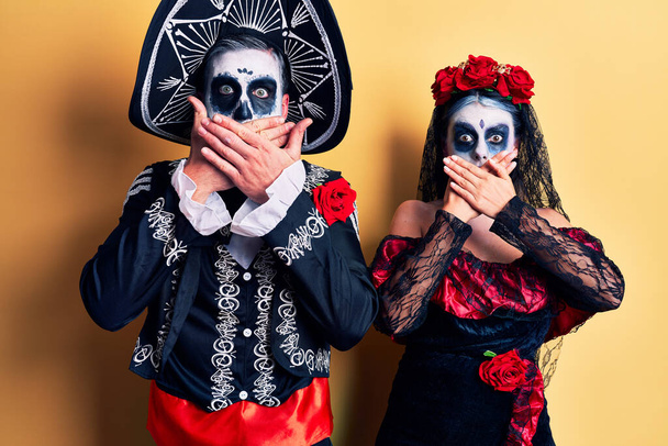 La coppia giovane che indossa il giorno messicano del costume morto sopra bocca di copertura scioccata gialla con mani per errore. concetto segreto.  - Foto, immagini