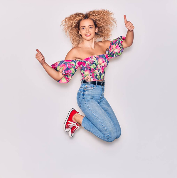 Young beautiful curly girl wearing casual clothes smiling happy. Jumping with smile on face doing ok sign with thumbs up over isolated white background. - Valokuva, kuva