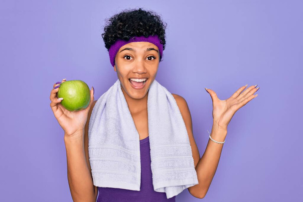 Mulher afro-americana africana bonita desportiva fazendo exercício vestindo toalha comendo maçã verde muito feliz e animado, expressão vencedora comemorando vitória gritando com grande sorriso e mãos levantadas
 - Foto, Imagem