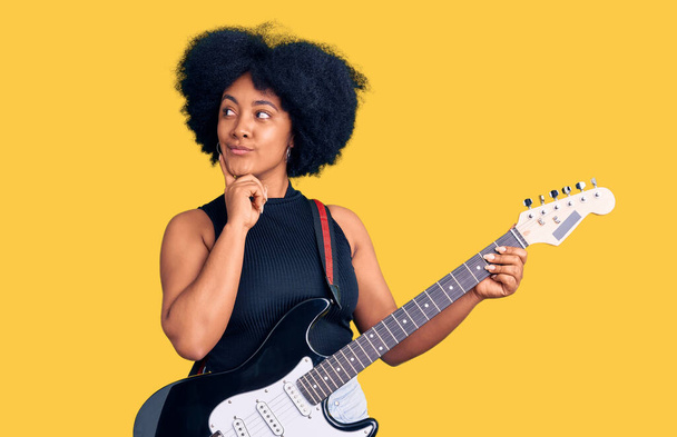 Jovem afro-americana tocando guitarra elétrica rosto sério pensando em questão com a mão no queixo, pensativo sobre ideia confusa
  - Foto, Imagem