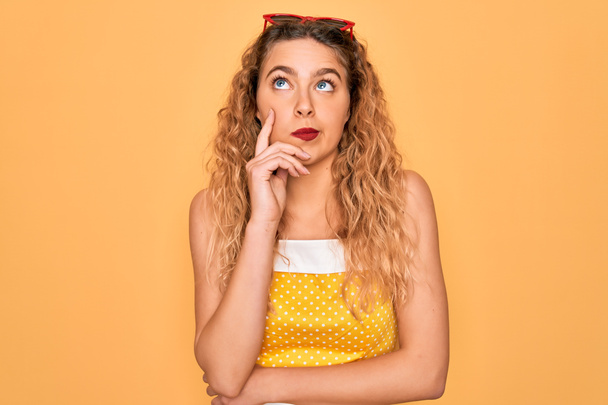 Beautiful blonde woman pin-up with blue eyes wearing red sunglasses over yellow background serious face thinking about question, very confused idea - Фото, зображення