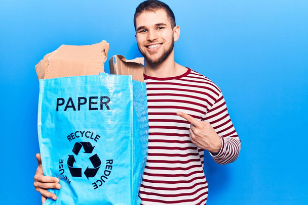 Молодой красивый мужчина перерабатывает бумагу улыбаясь счастливо указывая рукой и пальцем  - Фото, изображение