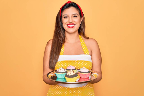 Young hispanic pin up woman wearing fashion sexy 50s style holding sweet cupcakes with a happy face standing and smiling with a confident smile showing teeth - Valokuva, kuva