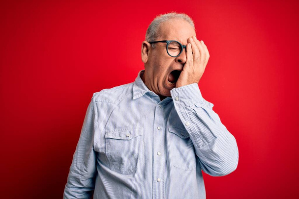 Moyen-âge beau homme satirique portant chemise rayée décontractée et des lunettes sur fond rouge bâillement fatigué couvrant demi-visage, oeil et bouche avec la main. Le visage souffre dans la douleur. - Photo, image