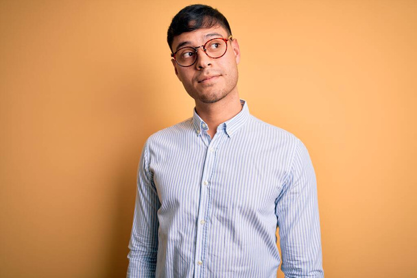 Junger gutaussehender hispanischer Geschäftsmann mit Nerd-Brille auf gelbem Hintergrund, lächelnd zur Seite schauend und nachdenkend wegstarrend. - Foto, Bild