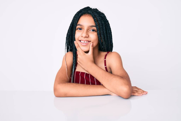 Mladá africká americká dívka s copánky na sobě ležérní oblečení sedí na stole a vypadá sebevědomě na kameru usmívá se zkříženýma rukama a rukou zvednutou na bradě. pozitivní myšlení.  - Fotografie, Obrázek