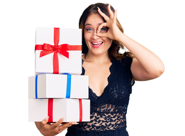 Νεαρή όμορφη καυκάσια γυναίκα κρατώντας δώρα χαμογελώντας ευτυχισμένη κάνει εντάξει σημάδι με το χέρι στο μάτι κοιτάζοντας μέσα από τα δάχτυλα  - Φωτογραφία, εικόνα