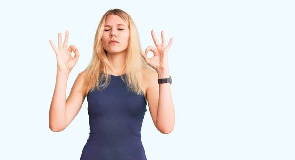 Jeune belle femme blonde vêtue d'une robe décontractée se détendre et sourire les yeux fermés en faisant geste de méditation avec les doigts. concept de yoga.  - Photo, image