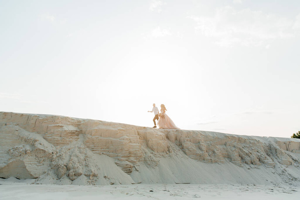 Histoire d'amour d'un beau couple dans une robe de luxe de mariage rose avec un bouquet dans le désert du Sahara, sable, dunes - Photo, image