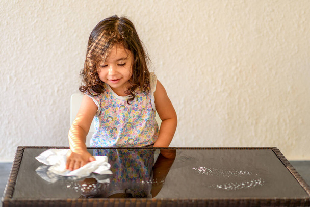 Schattig kind helpen moeder schoonmaken glazen tafel. Meisje maken schoonmaken in kamer thuis. - Foto, afbeelding