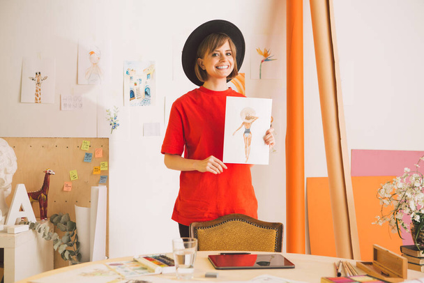 Pretty smiling girl artist in hat stands with drawing in art studio. Photography for ad of art courses or blog - 写真・画像