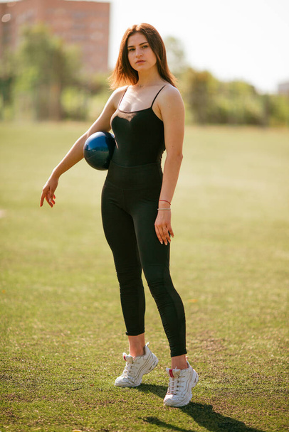 νέα όμορφη κοπέλα σε μαύρο αθλητικό ντύσιμο κάνει γυμναστική στις σχισμές στο πάρκο με μπάλα - Φωτογραφία, εικόνα