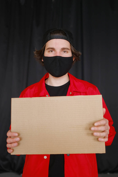 A young man of 25-30 years old in a black protective mask, yellow glasses, a cap and a red jacket holds an empty cardboard sign in his hands. - Fotografie, Obrázek