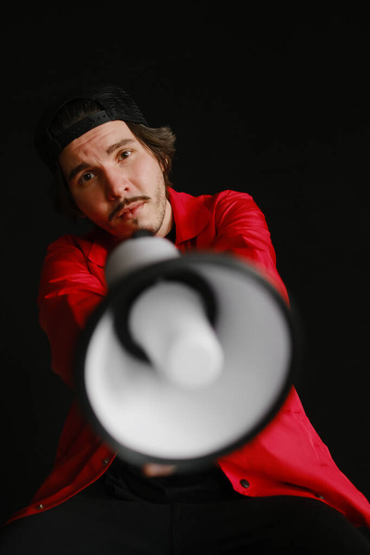 An emotional young man of 25-30 years old in a black cap and in a red jacket sits on a blue barrel and holds a megaphone in his hands on a dark background. Protest concept - Fotografie, Obrázek