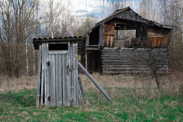 Vecchio villaggio di legno bagno squallido in primo piano con una casa abbandonata rotto sullo sfondo in campagna in primavera. Povertà nel villaggio russo - Foto, immagini