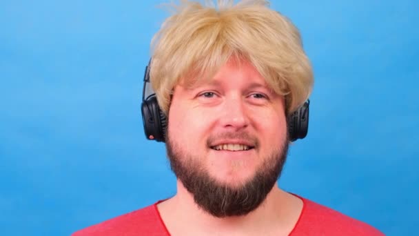 riendo hombre gordo raro en una peluca y una camiseta rosa en sus manos escucha música en los auriculares y baila sobre un fondo azul - Metraje, vídeo