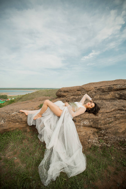 hermosa joven hembra con maquillaje y peinado en una boda suave peignoir se encuentra en las piedras al aire libre
 - Foto, Imagen