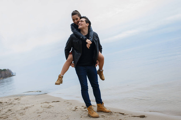 黒のジャケットを着た若いカップルがビーチで抱き合ってポーズをとって. - 写真・画像