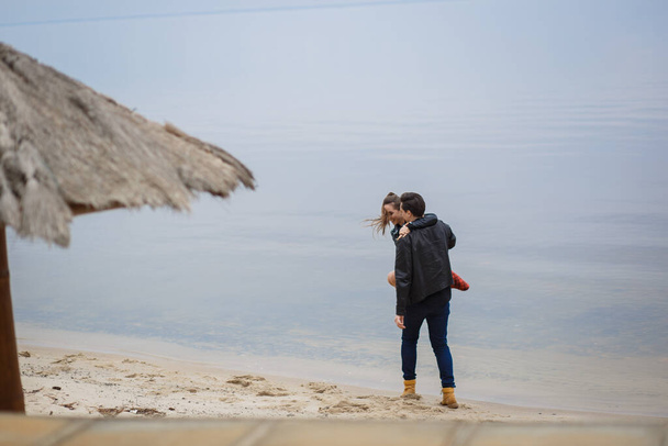 giovane coppia in giacche nere che si abbraccia e posa sulla spiaggia. - Foto, immagini