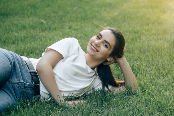 One young beautiful European brunette with dark eyes in a white T-shirt is lying on the grass holding her head and smiling. Caucasian woman laughing while lying on the lawn on a summer day. Place for text. - Foto, Imagem