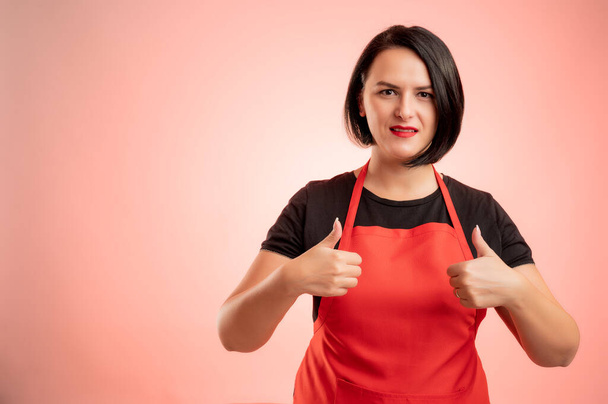Frau im Supermarkt mit roter Schürze und schwarzem T-Shirt, Daumen hoch, beide Hände isoliert auf rotem Hintergrund - Foto, Bild