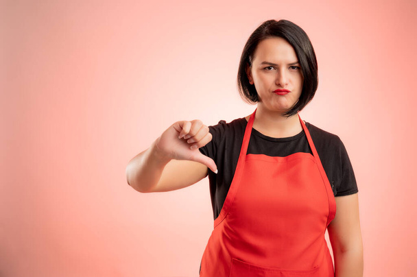Жінка, що працює в супермаркеті з червоним фартухом і чорною футболкою, демонструє нелюбов ізольовано на червоному тлі
 - Фото, зображення