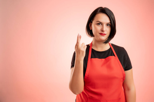 Γυναίκα που εργάζονται σε σούπερ μάρκετ με κόκκινο apronn και μαύρο t-shirt, δείχνουν καλή τύχη απομονώνονται σε κόκκινο φόντο - Φωτογραφία, εικόνα