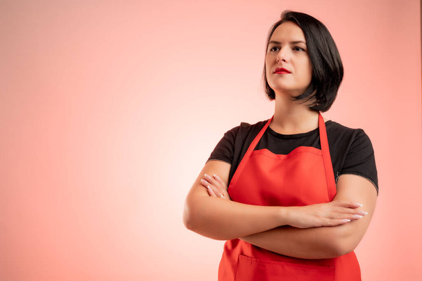 Woman employed at supermarket with red apron and black t-shirt, confident hero-shot isolated on red background - Photo, Image