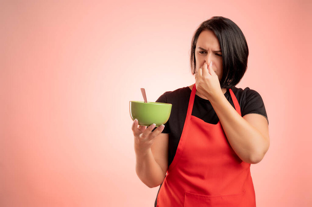 Woman employed at supermarket with red apron and black t-shirt, holding a bowl of soup that smells bad isolated on red background - Photo, Image