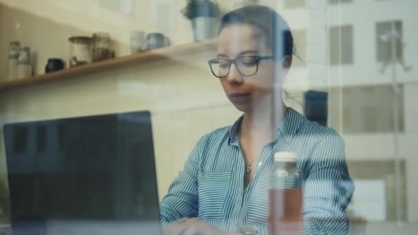 A woman operating a laptop is seen through a cafe window - Video, Çekim