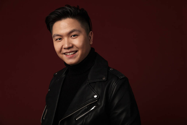 Image of joyful asian man in leather jacket smiling and looking at camera isolated over burgundy background - 写真・画像