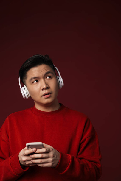 Image of thinking handsome asian man using headphones and cellphone isolated over burgundy background - Fotografie, Obrázek