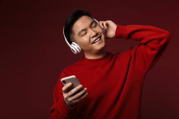 Image of joyful young asian man smiling while using headphones and cellphone isolated over burgundy background - Zdjęcie, obraz