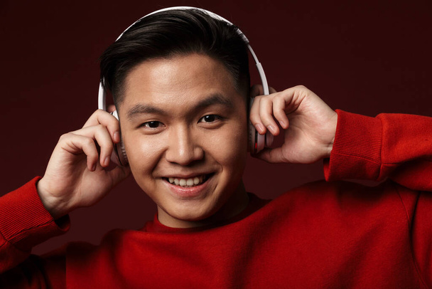 Image of joyful young asian man smiling while using wireless headphones isolated over burgundy background - 写真・画像
