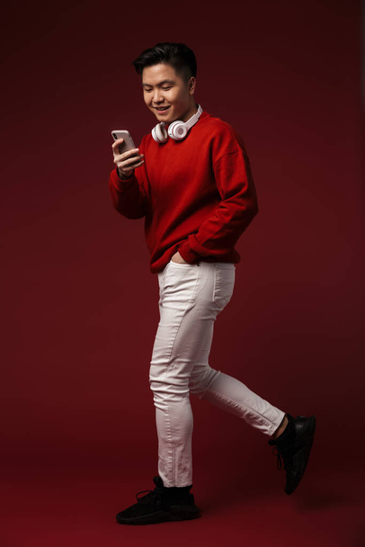 Obraz radosnego młodego azjatyckiego mężczyzny używającego słuchawek i telefonu komórkowego podczas spaceru wyizolowanego nad burgundowym tłem - Zdjęcie, obraz