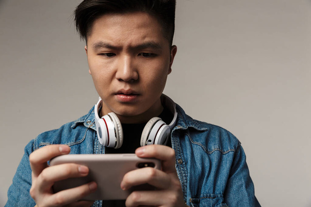 Image of focused young asian man playing video game on smartphone isolated over gray background - Photo, Image