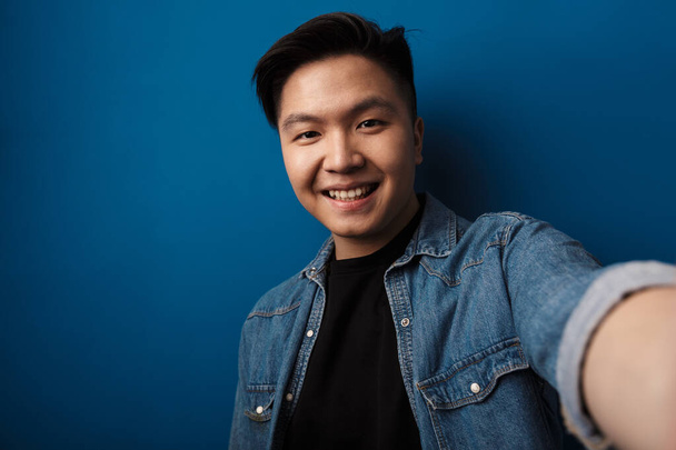 Image of joyful handsome asian man smiling and taking selfie photo isolated over blue background - Foto, Imagen
